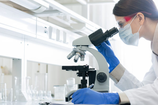 Young woman in whitecoat, gloves, eyewear and mask bending over microscope