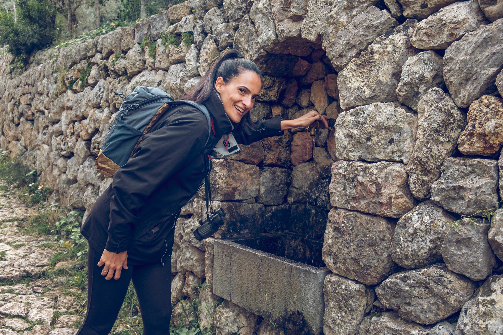 Positive female hiker standing near stone wall with flowing spring water and smiling