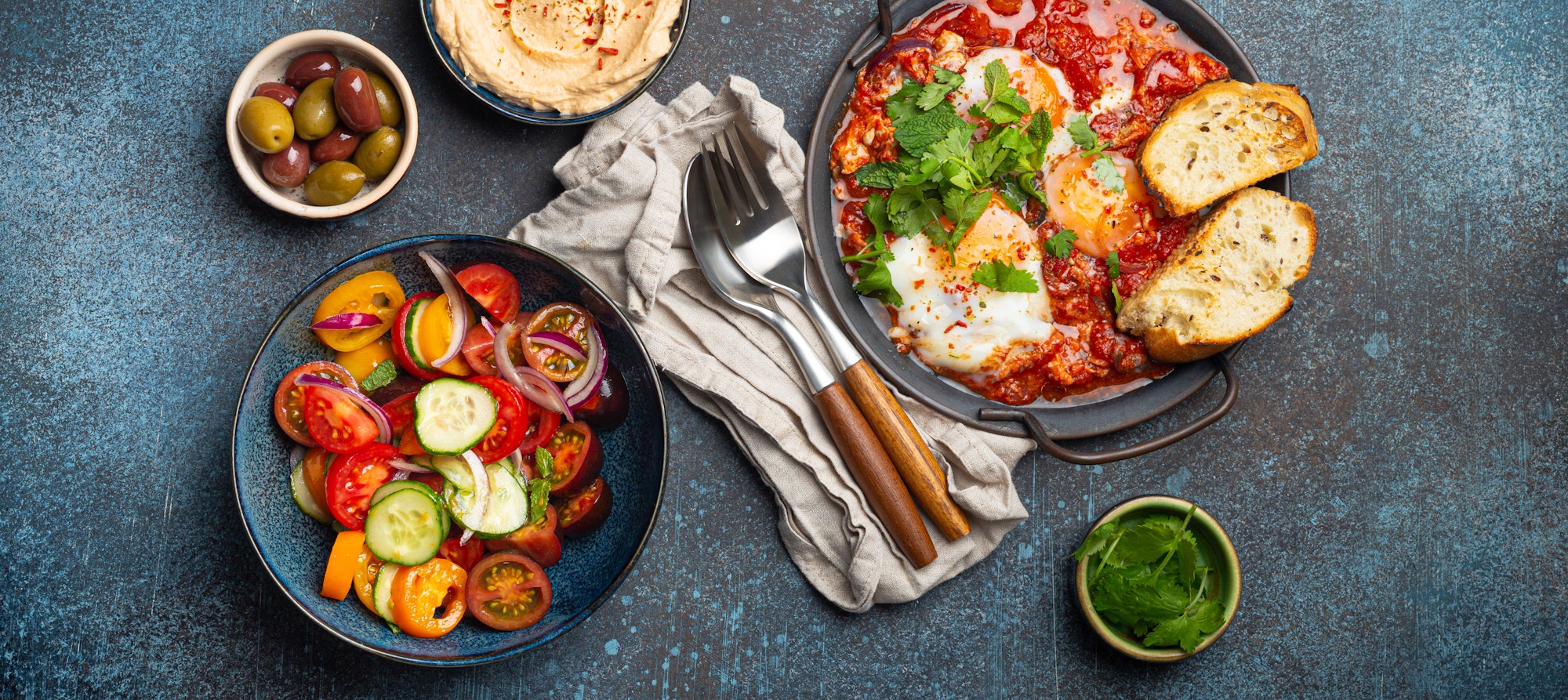 Middle Eastern breakfast or brunch with Shakshouka in pan, toasts, vegetables salad, hummus, olives