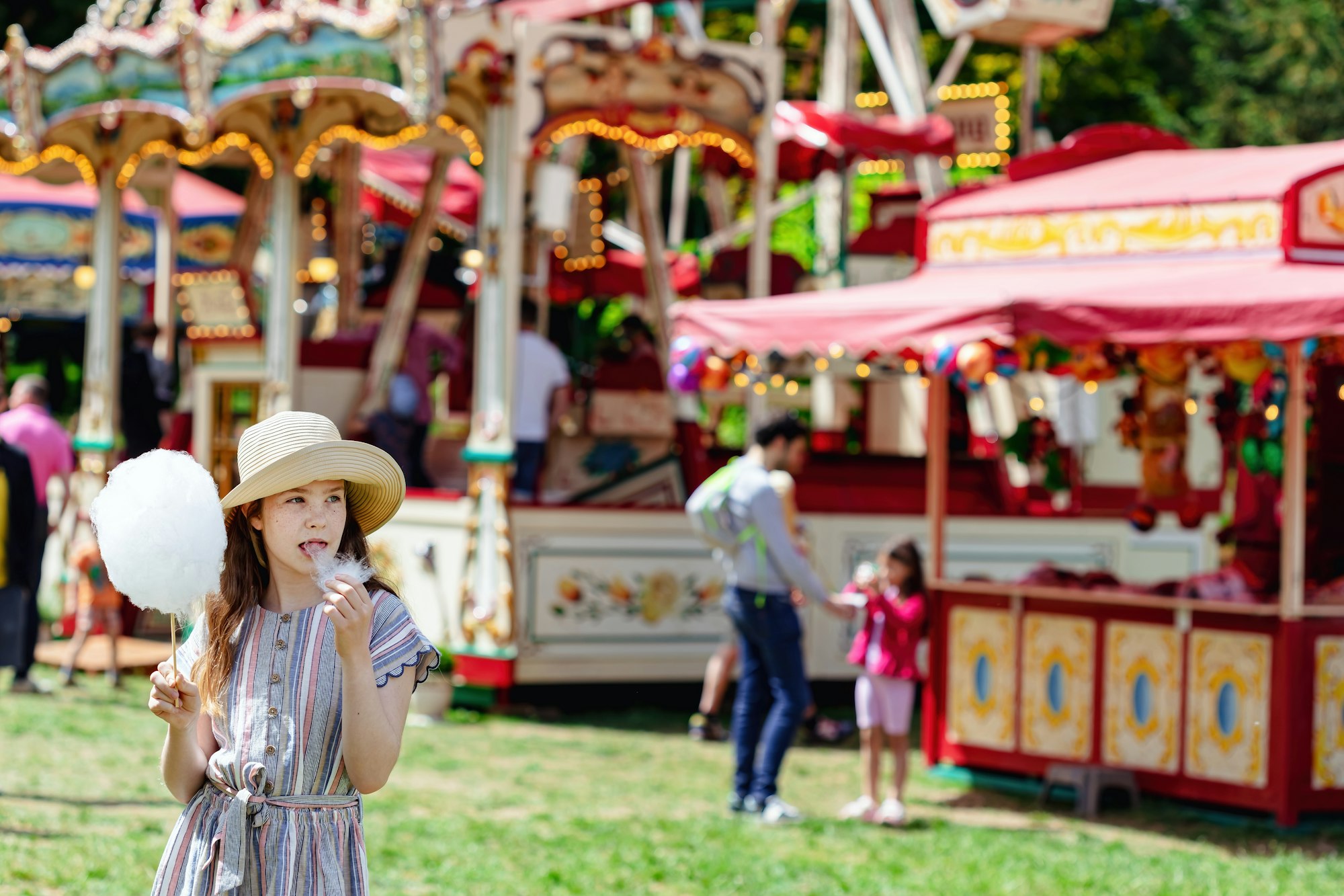 A girl in a hat and a dress eating cotton candy on a hot summer day at funfair