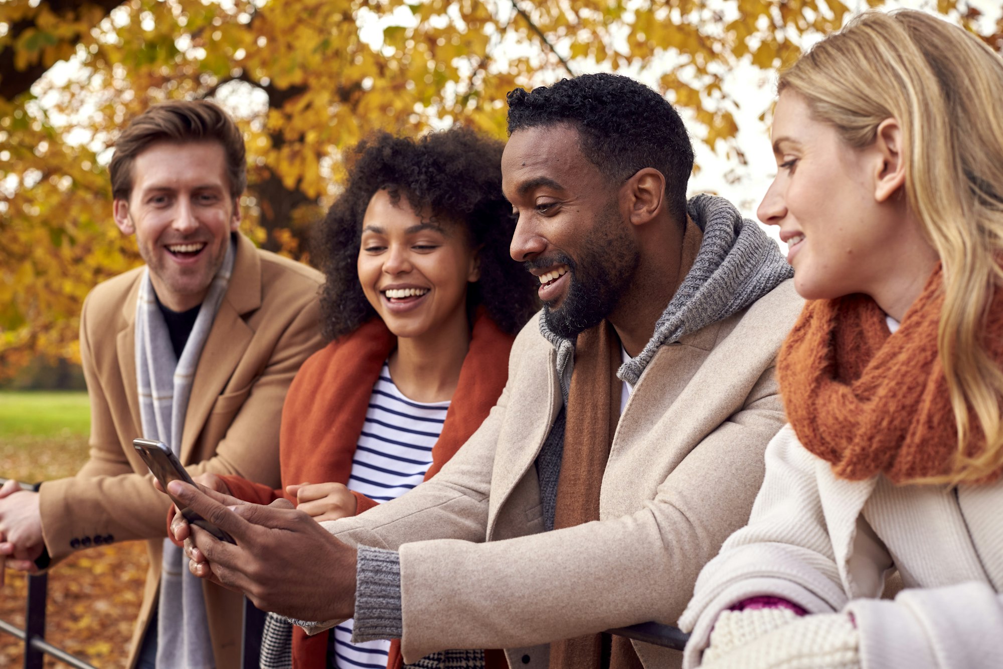 Group Of Friends Outdoors Wearing Coats And Scarves Looking At Photos On Phone In Autumn Park