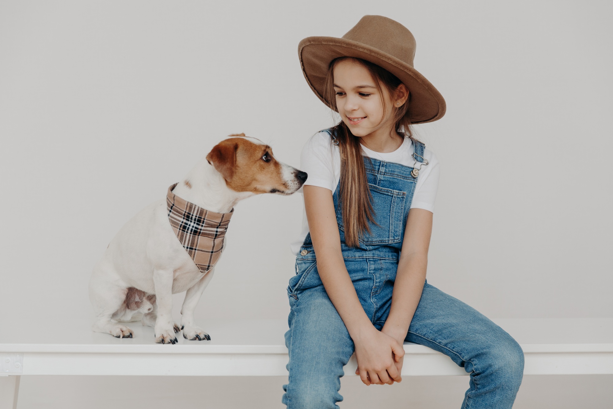 Isolated shot of pretty girl wears stylish hat, white t shirt and denim overalls, looks at her puppy Isolated shot of pretty girl wears stylish hat, white t shirt and denim overalls, looks at her puppy